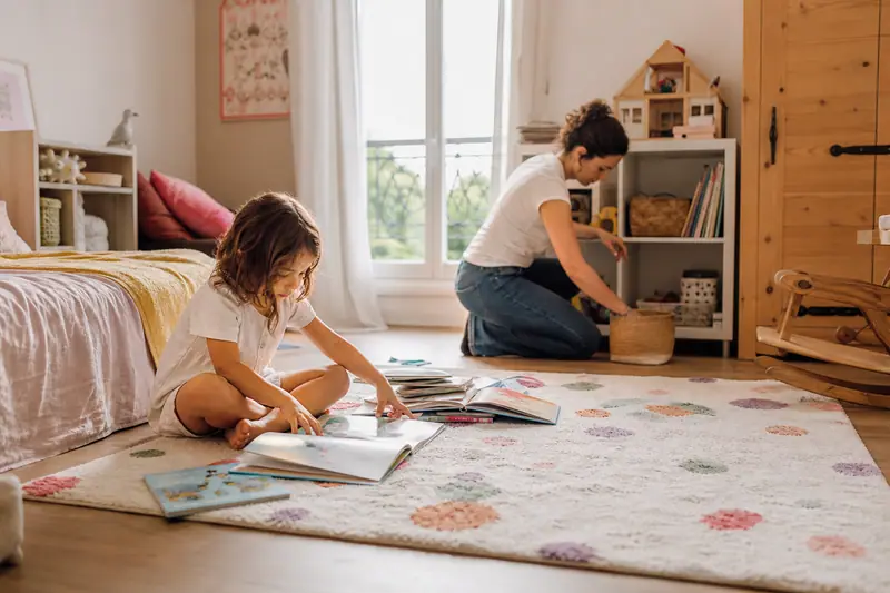 Un jeune enfant joue calmement avec des livres sur un tapis pendant que son parent range la chambre en arrière-plan.