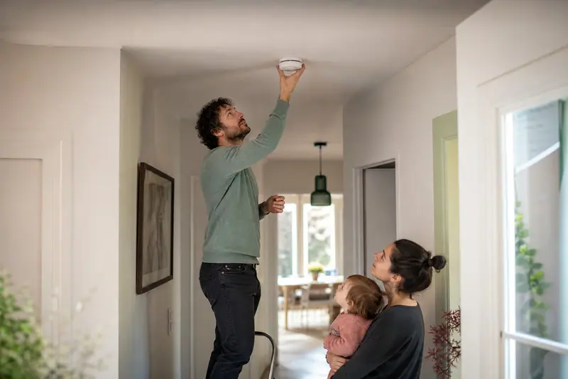 Parent debout sur un escabeau vérifiant un détecteur de fumée fixé au plafond d'un couloir familial.