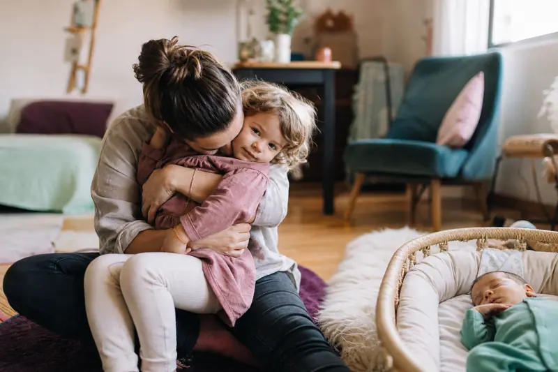 Parent assis au sol câlinant son enfant aîné pendant que le bébé dort dans un couffin à proximité, salon familial chaleureux.