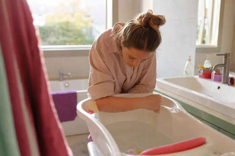 Parent testant la température de l'eau du bain avec le coude avant d'y installer un jeune enfant, salle de bain familiale lumineuse.