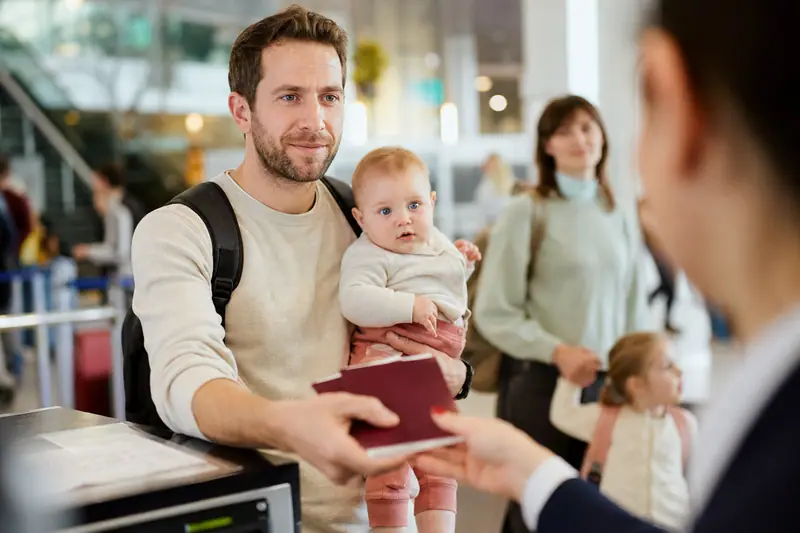 Parent tenant son bébé dans les bras devant un comptoir d'enregistrement d'aéroport, passeports et documents à la main.